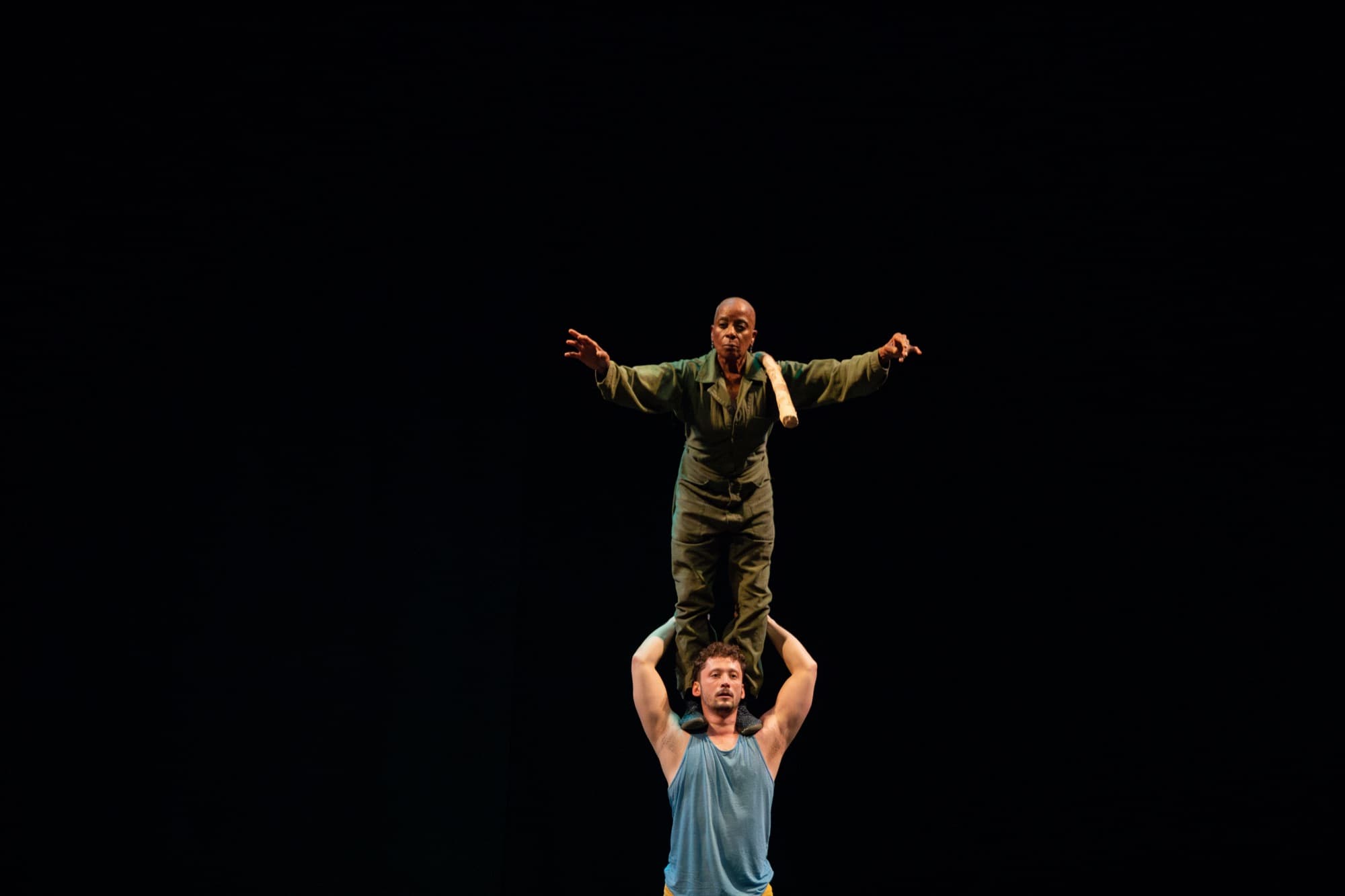 Rhonda stands on Ben's shoulders under a single spotlight.